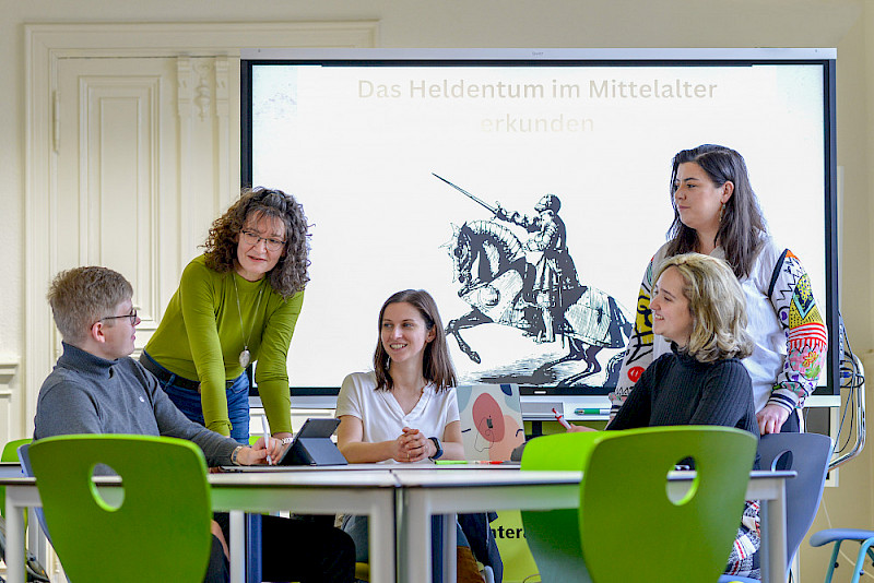Im Gespräch über das Unterrichtsmaterial, dessen Deckblatt auf dem Whiteboard zu sehen ist. Michael Beau, die Dozentinnen Andrea Seidel und Stefanie Naumann sowie Petya Vasileva (stehend) und Alona Polubotko (von links).